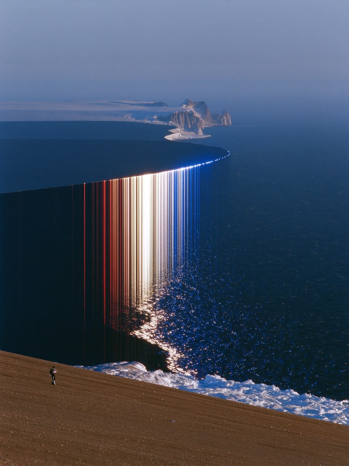 A figure stands above a vast ice shelf where a band of light pours down into the sea