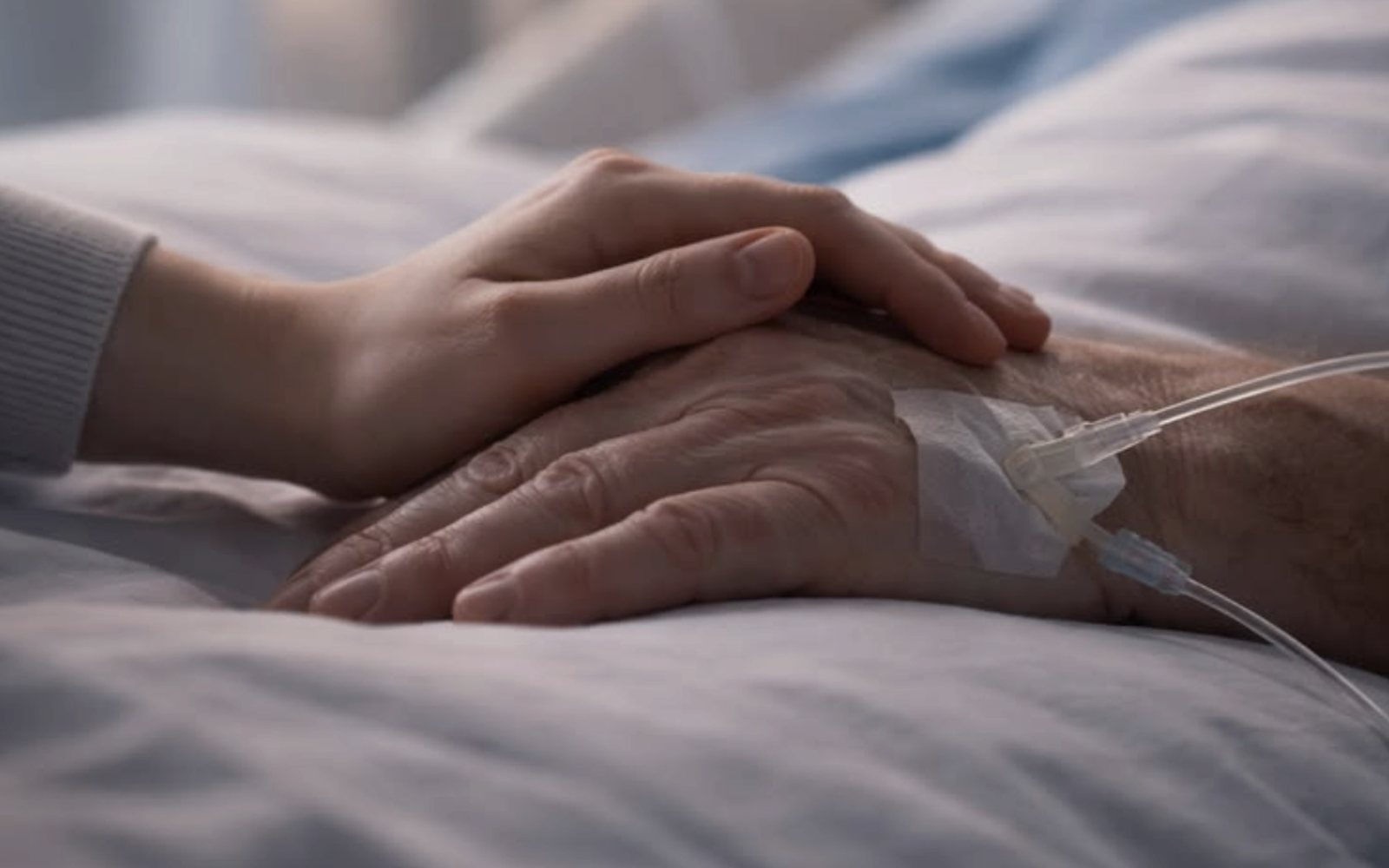 A hand resting on a patient’s hand beside a hospital bed