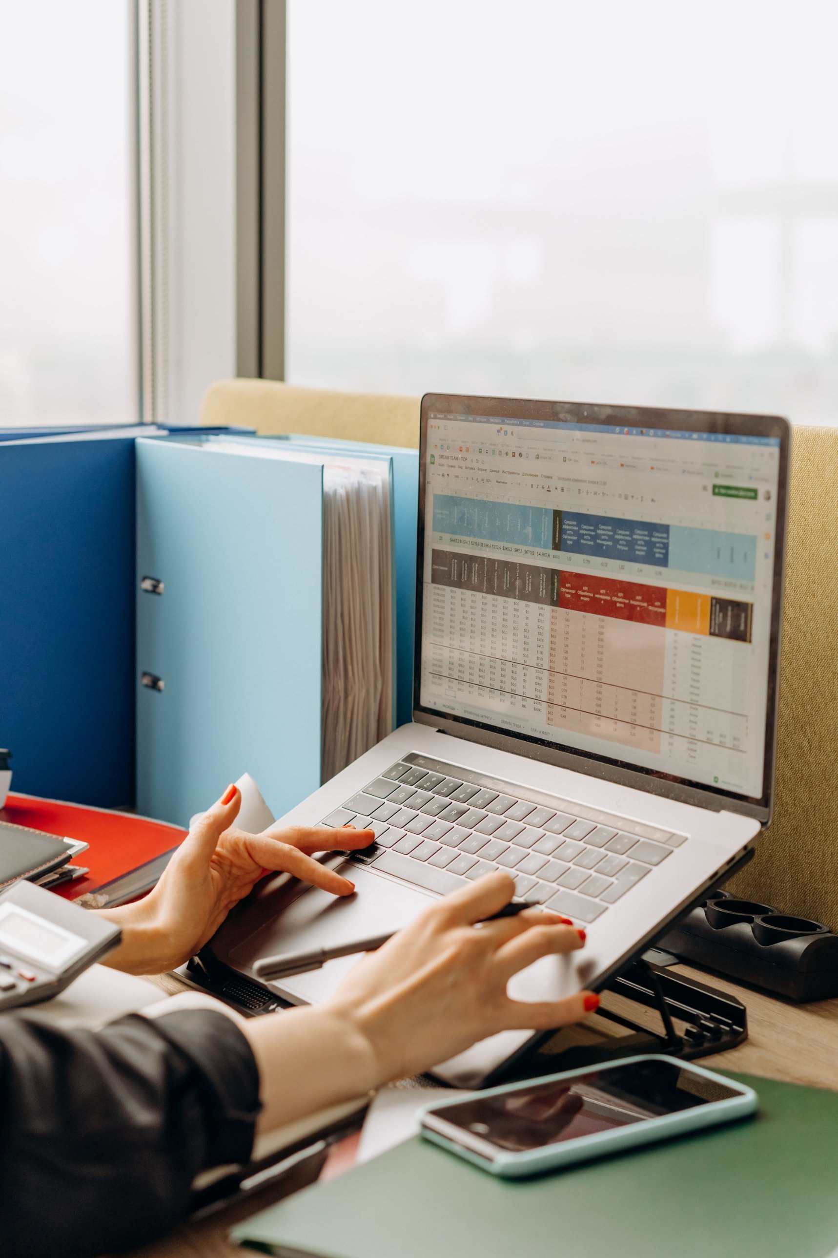 Hands working through a spreadsheet on a laptop beside stacked ring binders